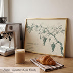 a framed print of a vineyard scene with grapes and leaves, a croissant, a cup of coffee, and a coffee maker on a wooden countertop.