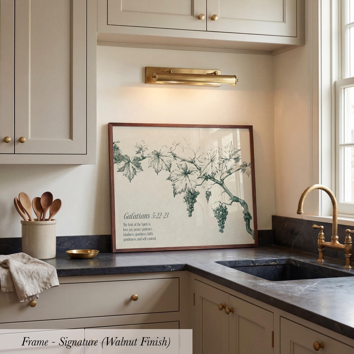 a kitchen counter with a framed print of a vineyard scene, a bowl of wooden spoons, and a sink.