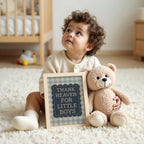 A young child sits on the floor, holding a framed sign that says "THANK HEAVEN FOR LITTLE BOYS" and a teddy bear.