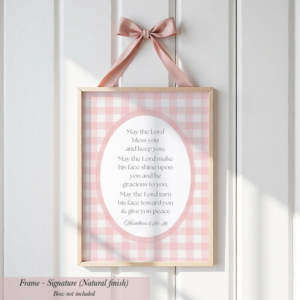 A framed sign with a pink and white checkered background hangs on a white wall. The sign contains a religious message, and the frame is described as a "Signature (Natural finish)" without further details.