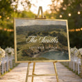 a wedding ceremony setup with a large sign displaying the names of the couple and the date of their wedding, surrounded by white chairs and a scenic outdoor setting with trees and string lights.