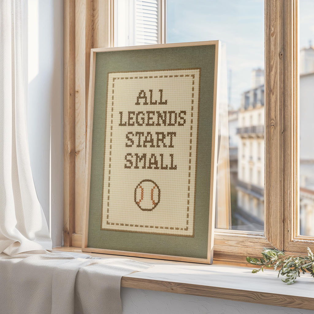 A framed cross-stitch design with the text "ALL LEGENDS START SMALL" and a baseball motif, placed on a windowsill.