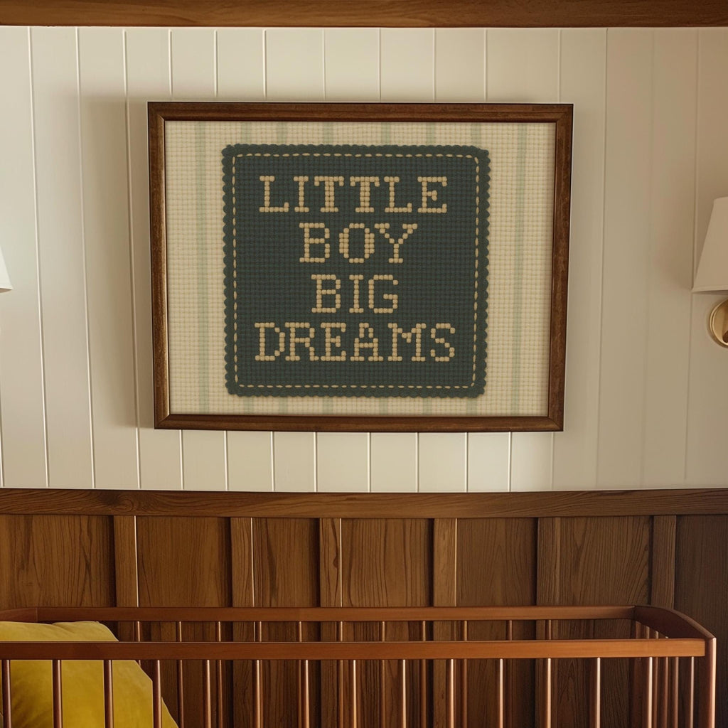a framed cross-stitch design with the text "LITTLE BOY BIG DREAMS" on it, hanging on a wall above a wooden crib.