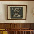 a framed cross-stitch design with the text "LITTLE BOY BIG DREAMS" on it, hanging on a wall above a wooden crib.