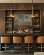 a modern bar area with a wooden wall, a large framed picture, and a variety of bottles and glasses displayed on the shelves. There are four bar stools in front of the bar, and two hanging pendant lights above the bar.