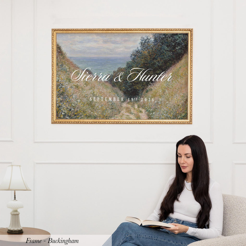 A woman is sitting on a couch, reading a book, with a framed painting of a landscape hanging on the wall above her.