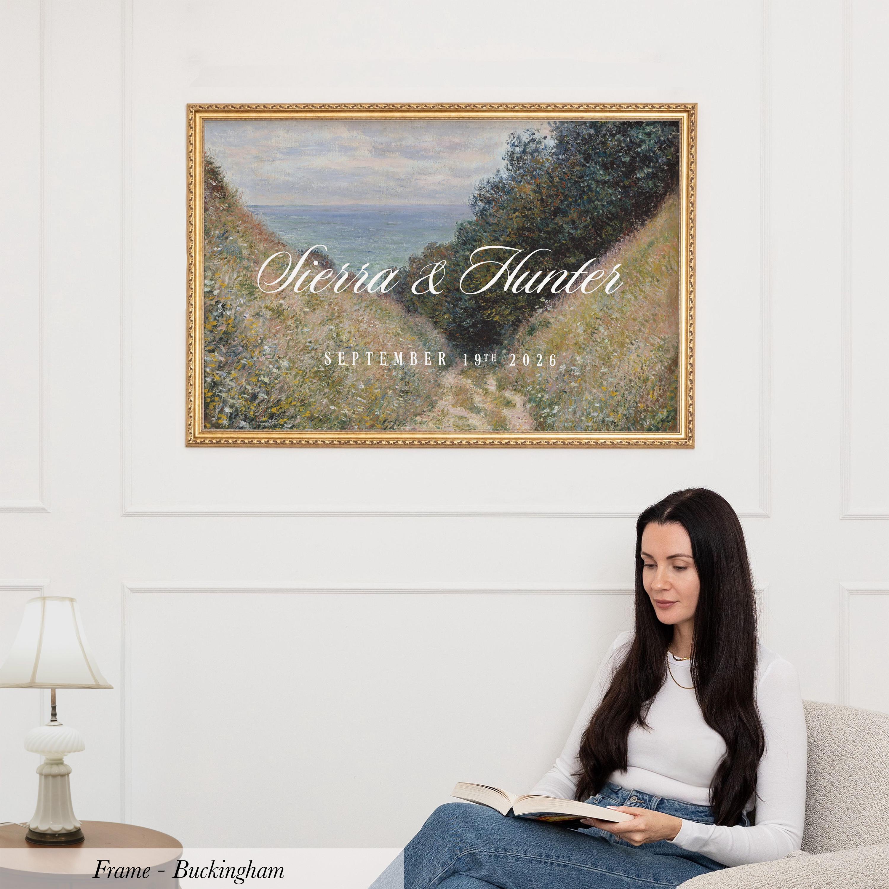 A woman is sitting on a couch, reading a book, with a framed painting of a landscape hanging on the wall above her.
