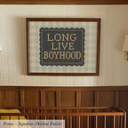 a framed cross-stitch design with the text "LONG LIVE BOYHOOD" on it, hanging on a wall above a wooden crib.