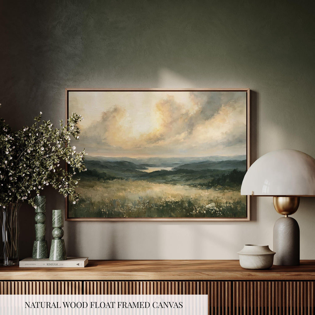 a wooden shelf with a framed landscape painting hanging on the wall above it. The painting depicts a serene countryside scene with rolling hills, a lake, and a cloudy sky. The shelf also includes a lamp, a vase with flowers, and a book, creating a cozy and inviting atmosphere.