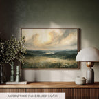 a wooden shelf with a framed landscape painting hanging on the wall above it. The painting depicts a serene countryside scene with rolling hills, a lake, and a cloudy sky. The shelf also includes a lamp, a vase with flowers, and a book, creating a cozy and inviting atmosphere.