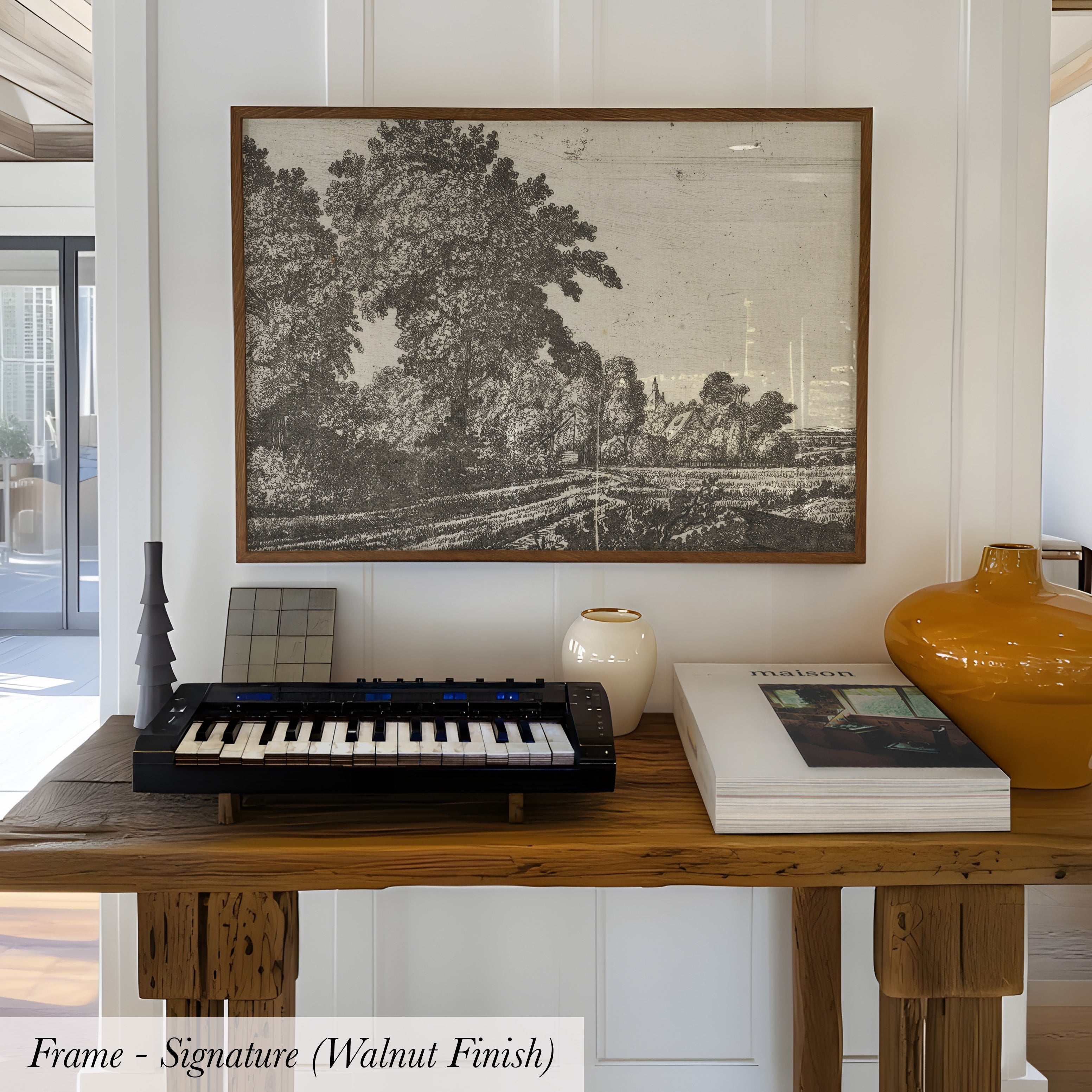 a wooden table with a framed black and white landscape painting hanging above it, a black and white keyboard, a white vase, and a stack of books.