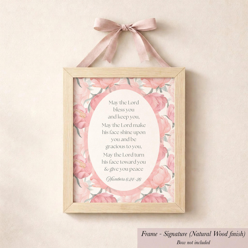 A framed sign with a pink floral design hangs on a wall, featuring a religious text.