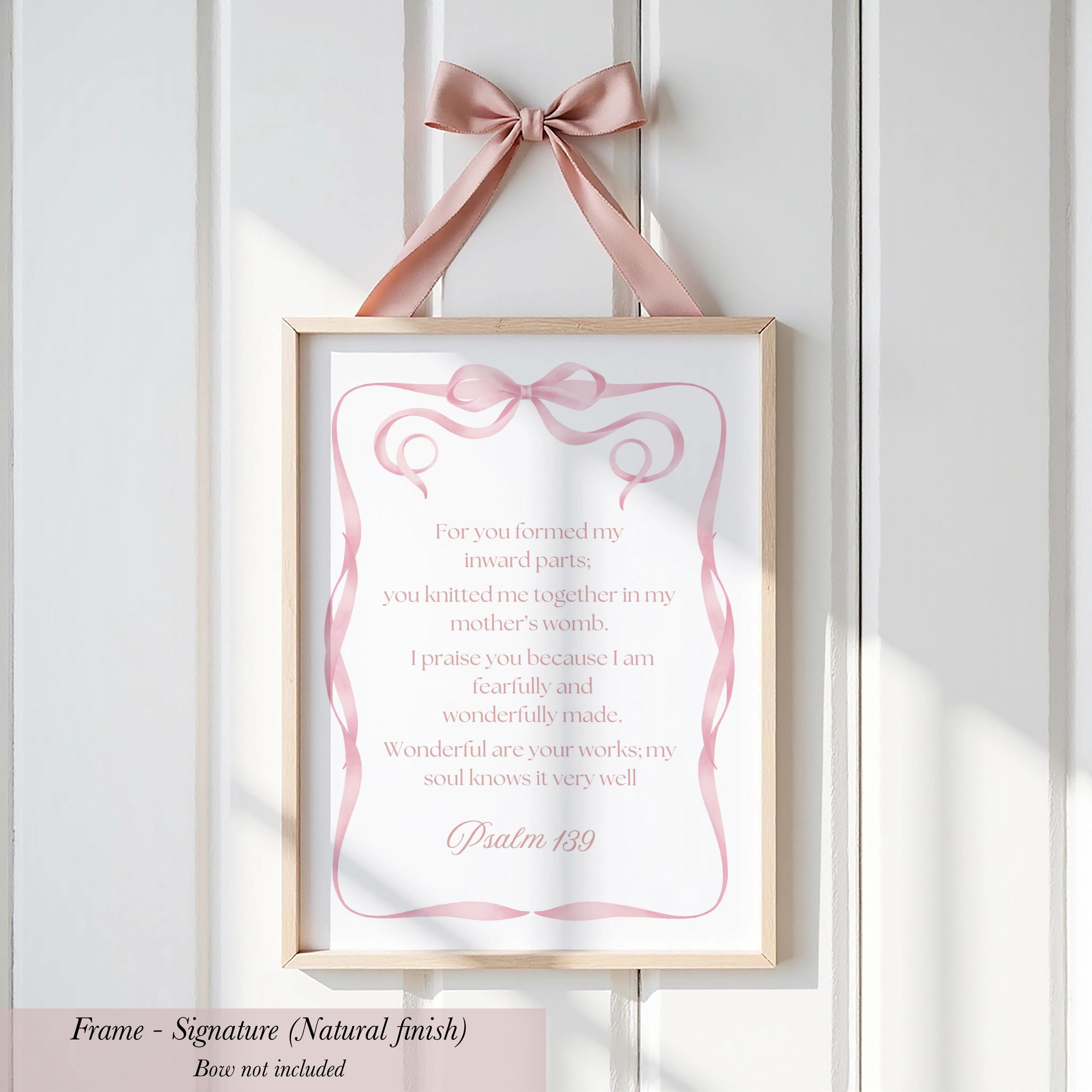 a framed sign with a pink ribbon bow, hanging on a white wall.