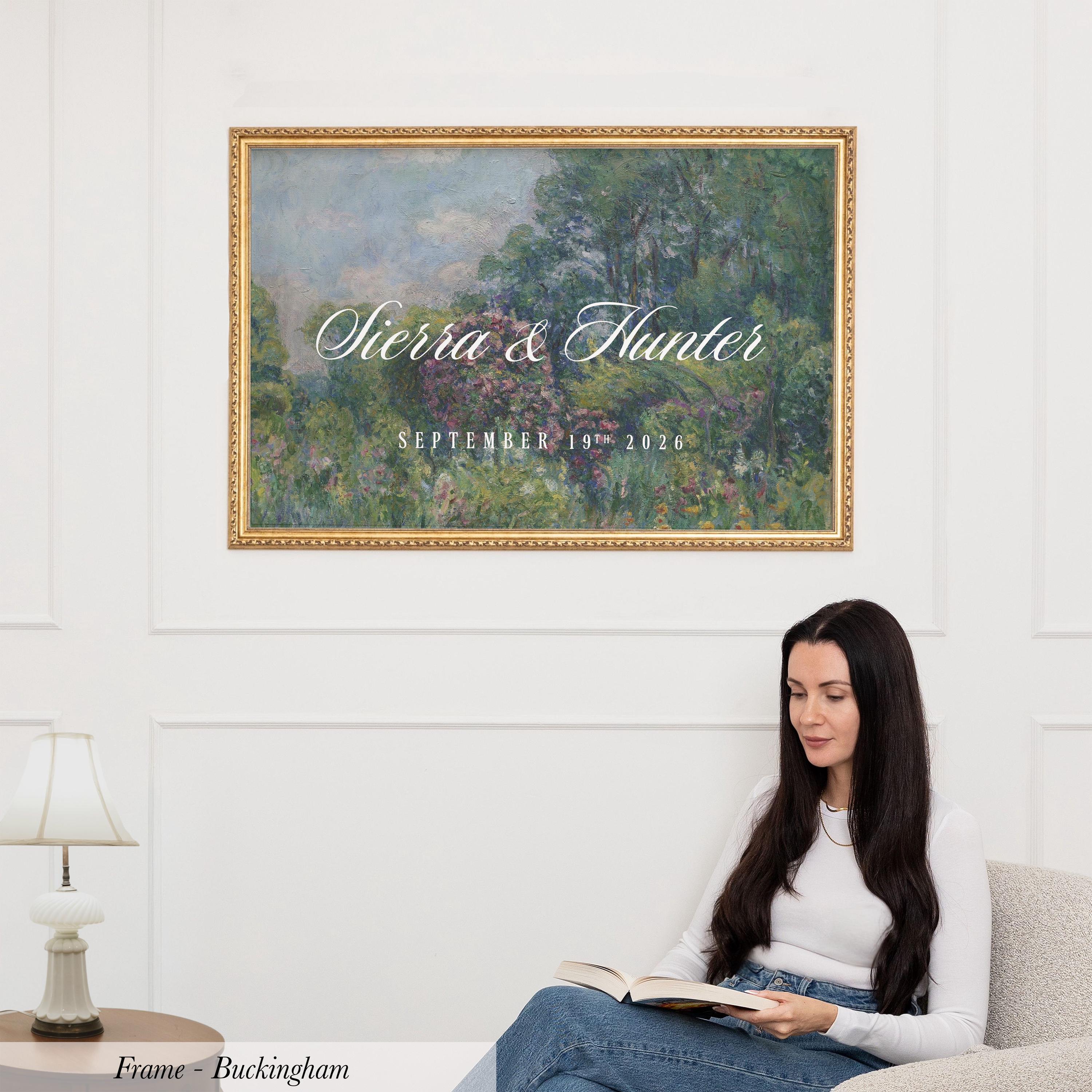 A woman is sitting on a couch, reading a book, with a framed painting of a landscape hanging on the wall behind her.
