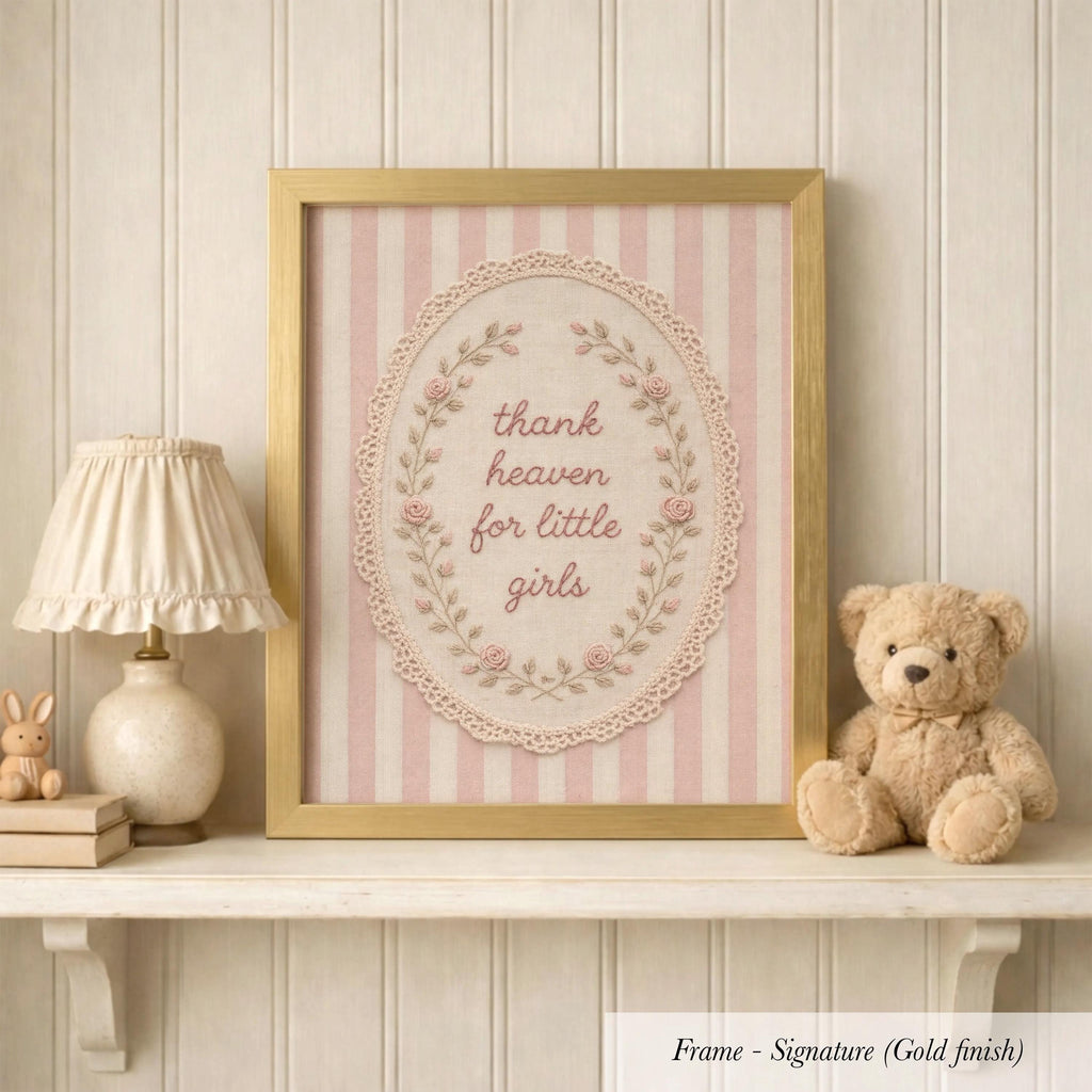 A framed picture with a pink and white striped border, displaying the text "Thank heaven for little girls" in a decorative floral design. The frame is placed on a shelf next to a lamp and a teddy bear.