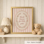 A framed picture with a pink and white striped border, displaying the text "Thank heaven for little girls" in a decorative floral design. The frame is placed on a shelf next to a lamp and a teddy bear.