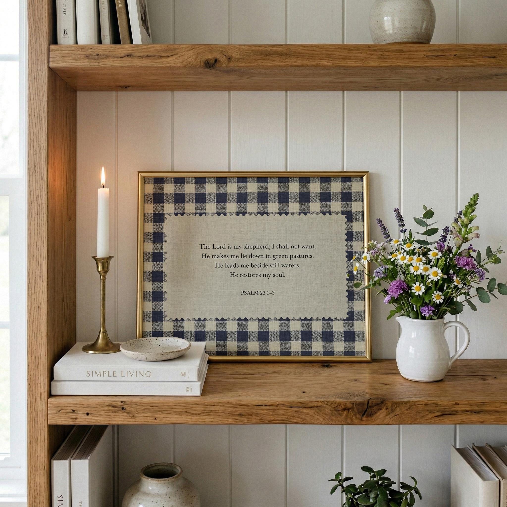 a wooden bookshelf with a framed Bible verse, a white candle, a white vase with flowers, and a stack of books.
