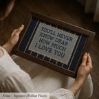 A person is holding a framed cross-stitch embroidery with the message "YOU'LL NEVER KNOW HOW DEAR I LOVE YOU" written on it.