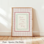 A framed print with a pink and white checkered border and the text "Thank Heaven for Little Girls" is displayed on a wooden floor against a white wall.