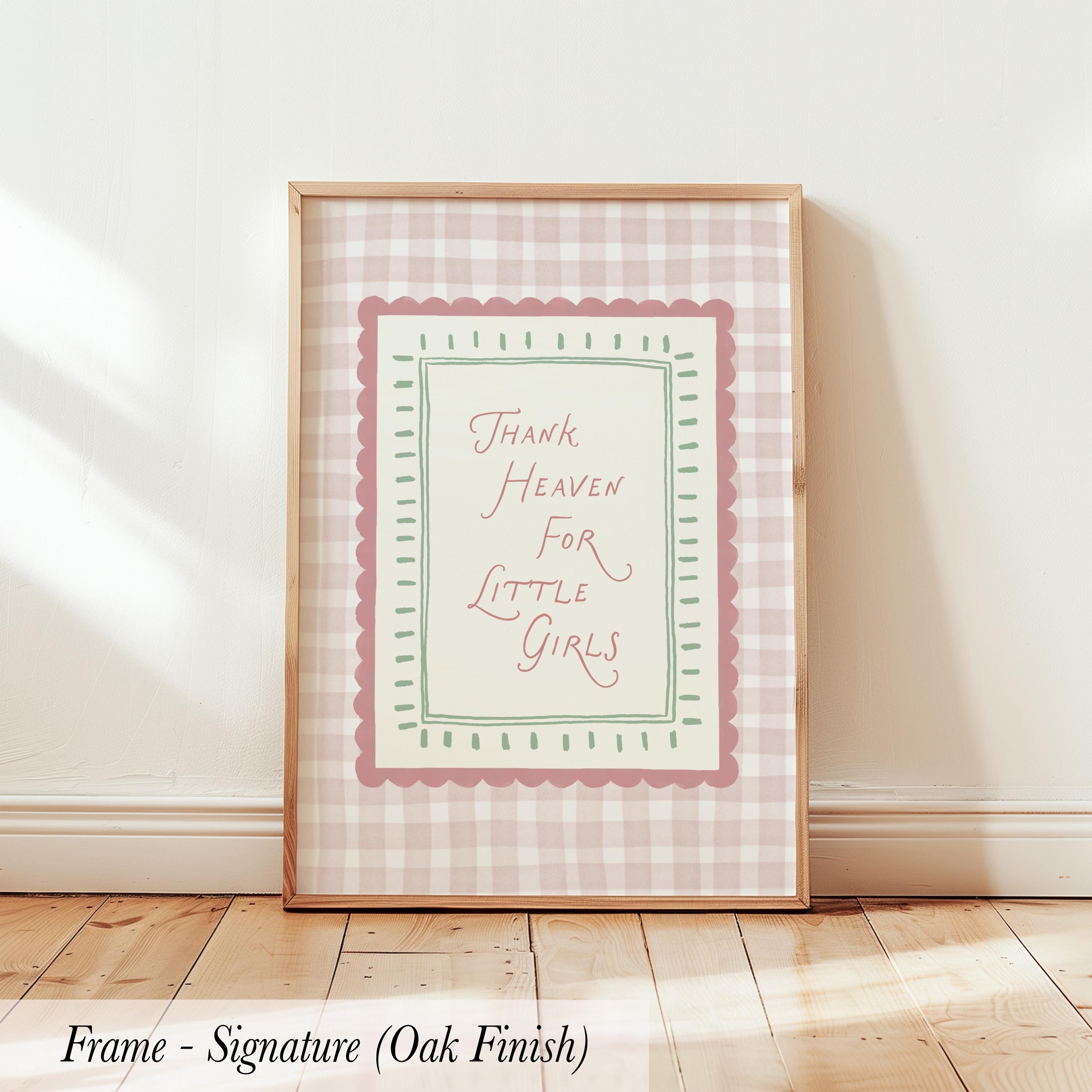 A framed print with a pink and white checkered border and the text "Thank Heaven for Little Girls" is displayed on a wooden floor against a white wall.