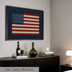 a framed American flag hanging on a wall above a black dresser with various decorative items on top, including a bottle, a vase, and a stack of books.