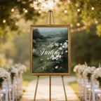 a wedding ceremony setup with a large framed sign displaying the names of the bride and groom, surrounded by white flowers and greenery. The sign is placed on a stone path, with rows of white chairs arranged in front of it. The scene is set against a backdrop of trees and string lights, creating a romantic and elegant atmosphere.