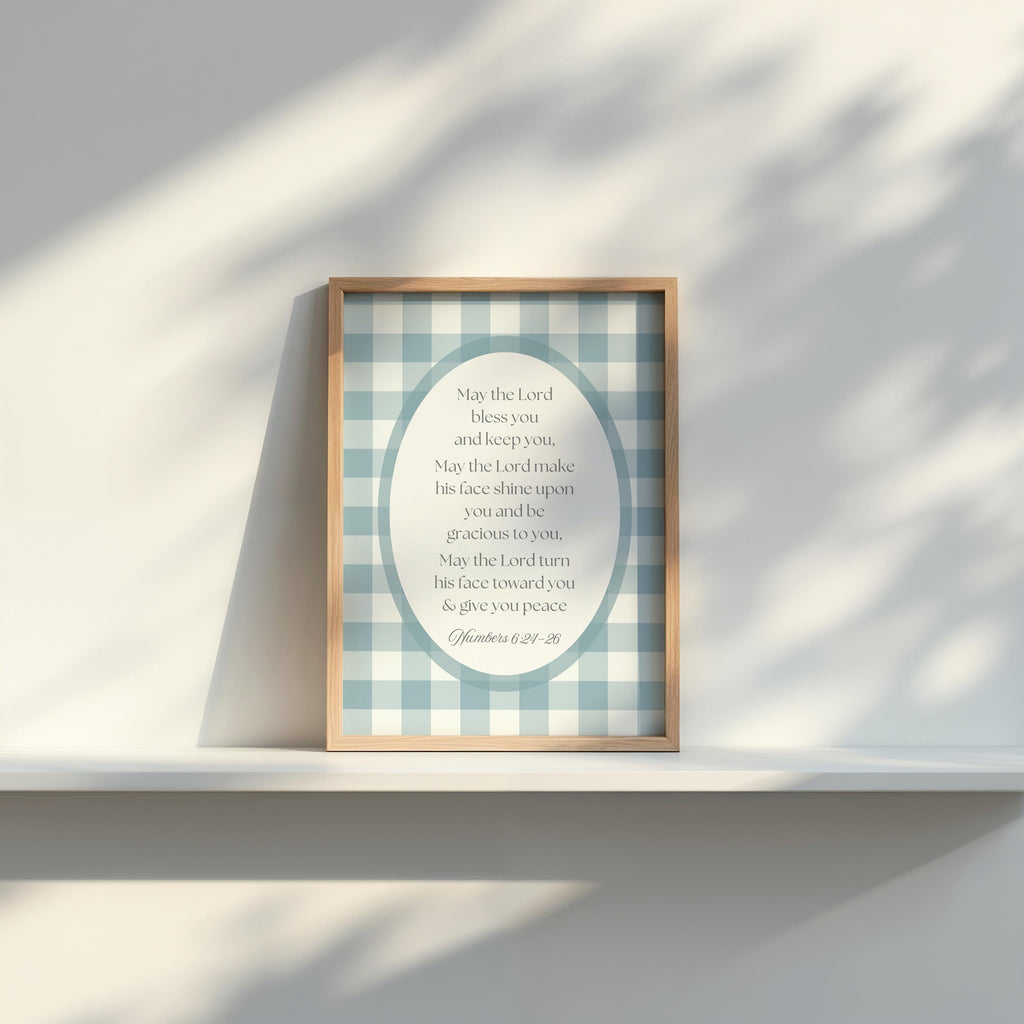 A framed print with a blue and white checkered background and a religious text is displayed on a white shelf against a white wall.
