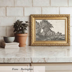 A framed black and white landscape painting is displayed on a marble countertop, accompanied by a potted plant, a stack of books, and a bowl.