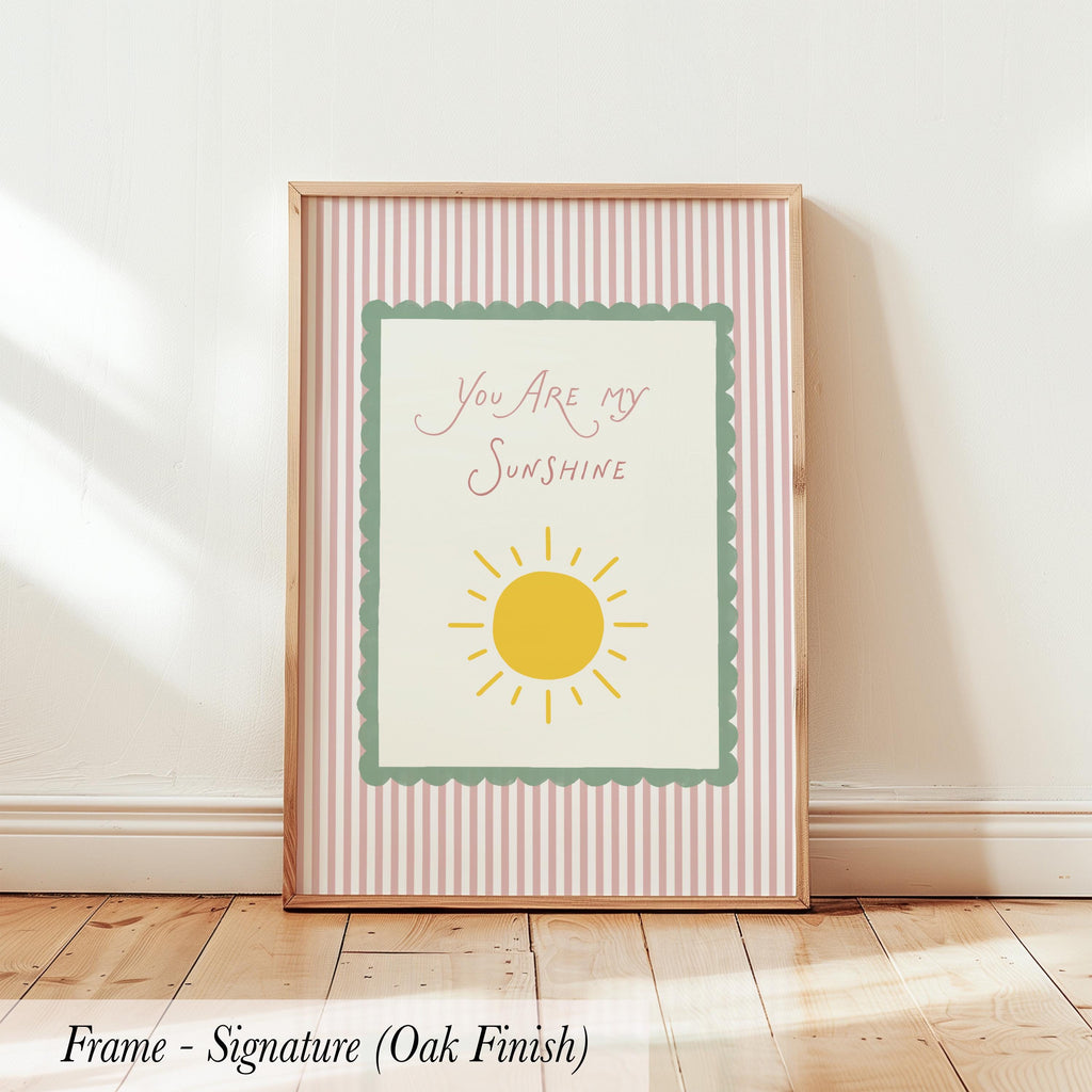 A framed poster with the text "You are my sunshine" and a sun graphic, placed on a wooden floor against a white wall.