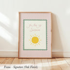A framed poster with the text "You are my sunshine" and a sun graphic, placed on a wooden floor against a white wall.