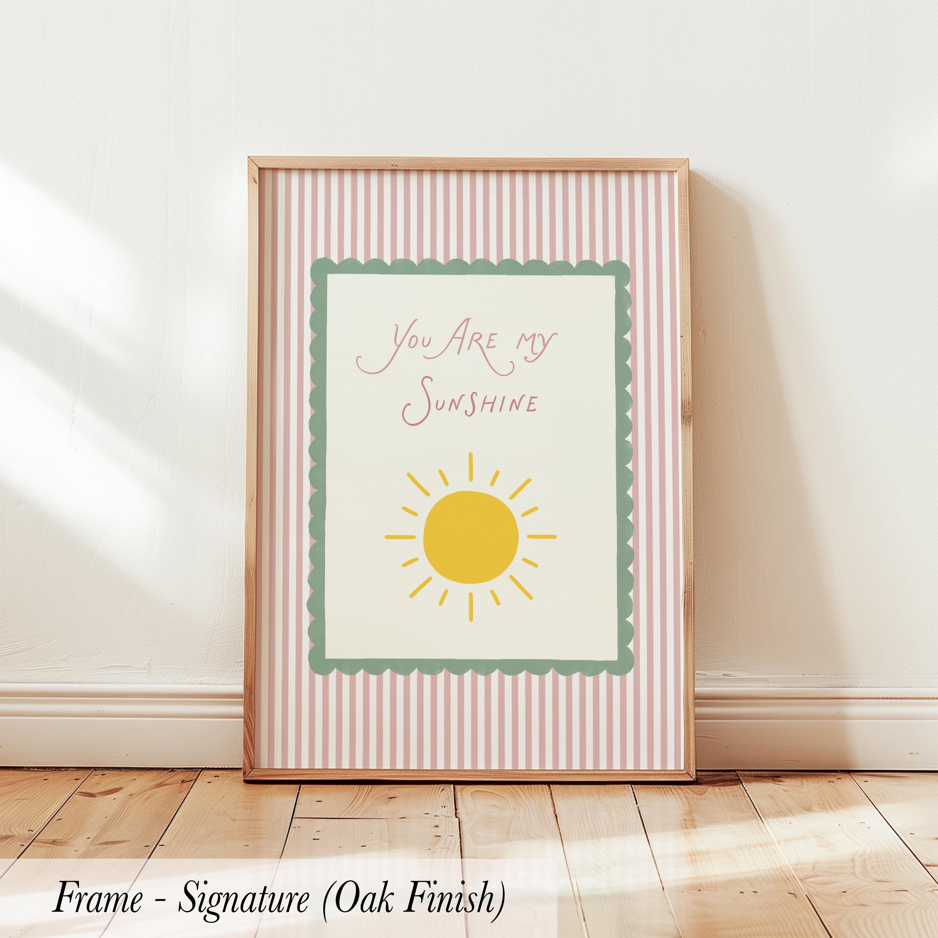 A framed poster with the text "You are my sunshine" and a sun graphic, placed on a wooden floor against a white wall.