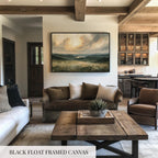 a living room with a large black-framed canvas painting hanging on the wall, depicting a scenic landscape. The room is furnished with a couch, a chair, and a coffee table, creating a cozy and inviting atmosphere.